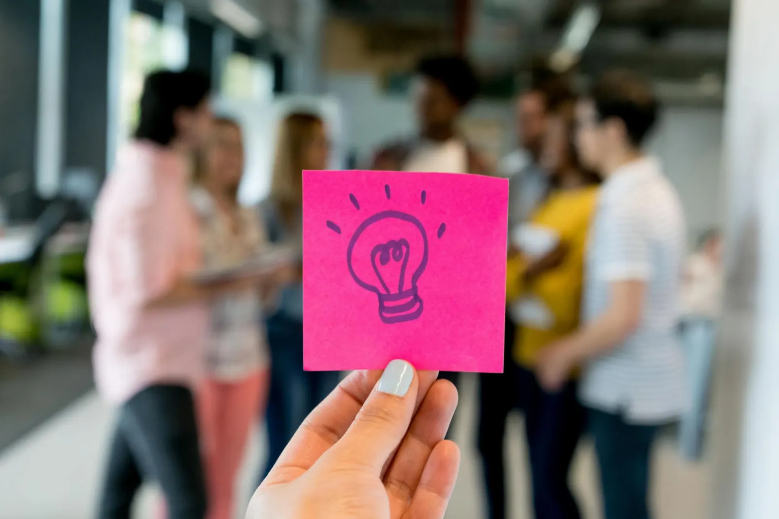 A hand holding a pink sticky note with a light-bulb sketch in front of a group of colleagues