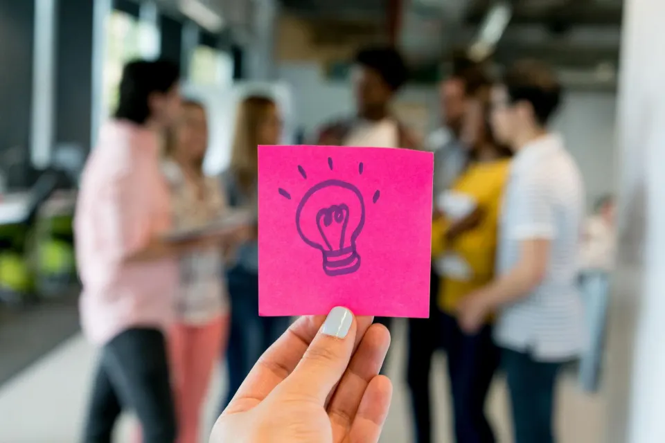 A hand holding a pink sticky note with a light-bulb sketch in front of a group of colleagues