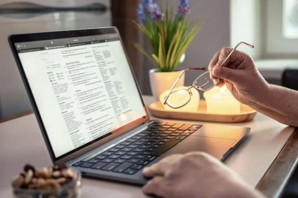 A person holding reading glasses while reading a document on a laptop